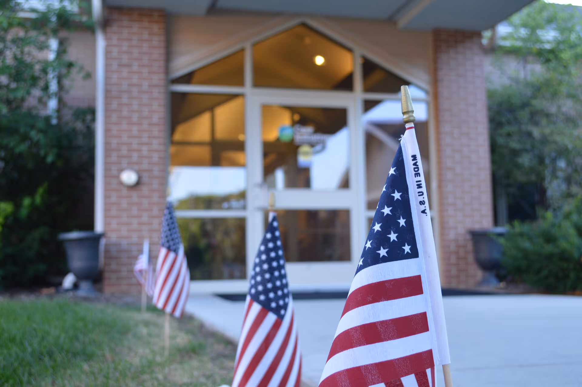 American flags lined up along a sidewalk leading to the entrance of a brick building with glass doors.