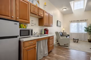 Interior view of a senior living facility room showing a small kitchen area with wooden cabinets, a mini refrigerator, microwave, and coffee maker. Adjacent to the kitchen is a living area with a recliner chair, a small round table, a couch, a floor lamp, and a large window with curtains allowing natural light to fill the space.