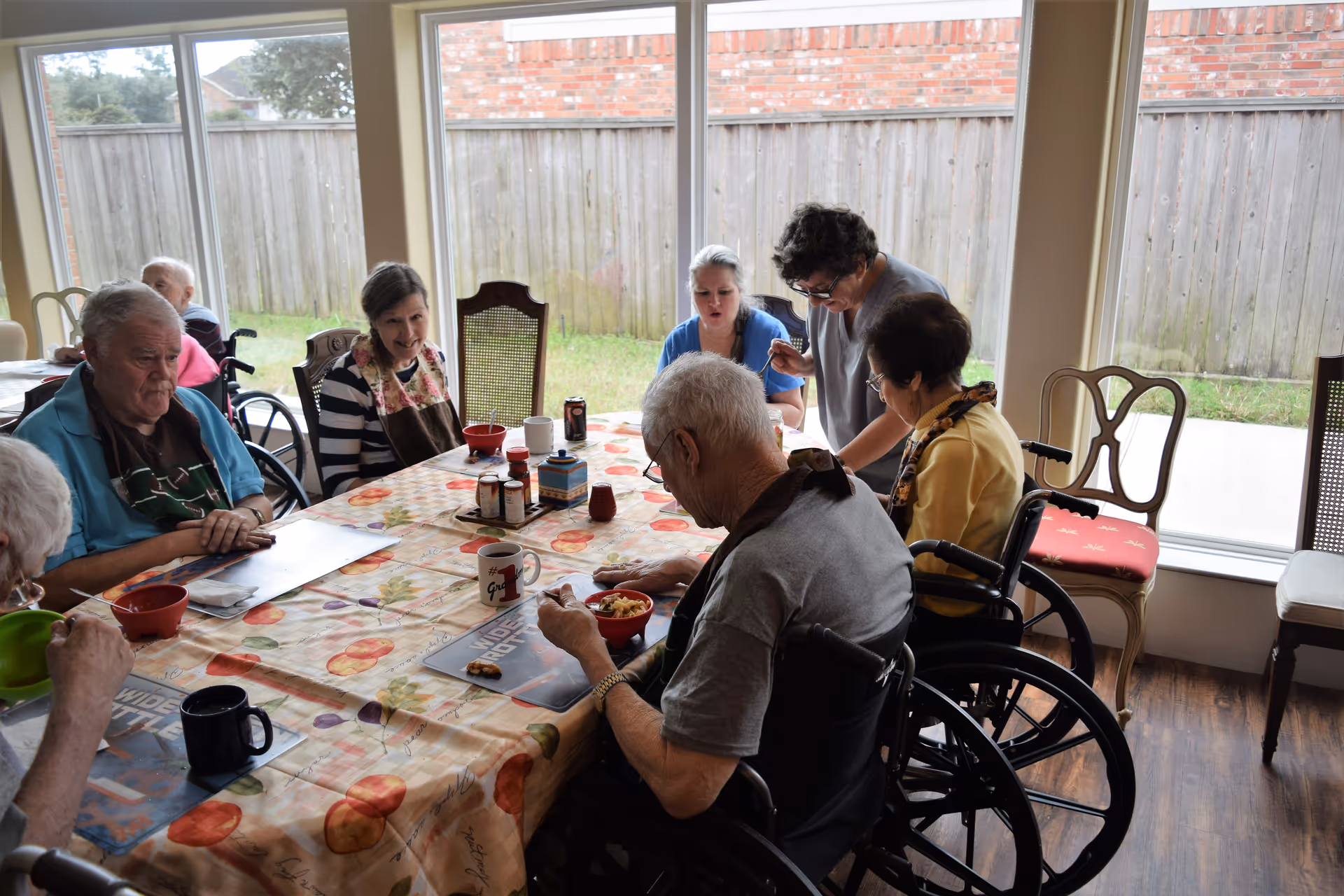 A group of elderly people, some in wheelchairs, sitting around a dining table covered with a colorful tablecloth. Two caregivers are assisting them during a meal in a bright room with large windows showing a fenced backyard.