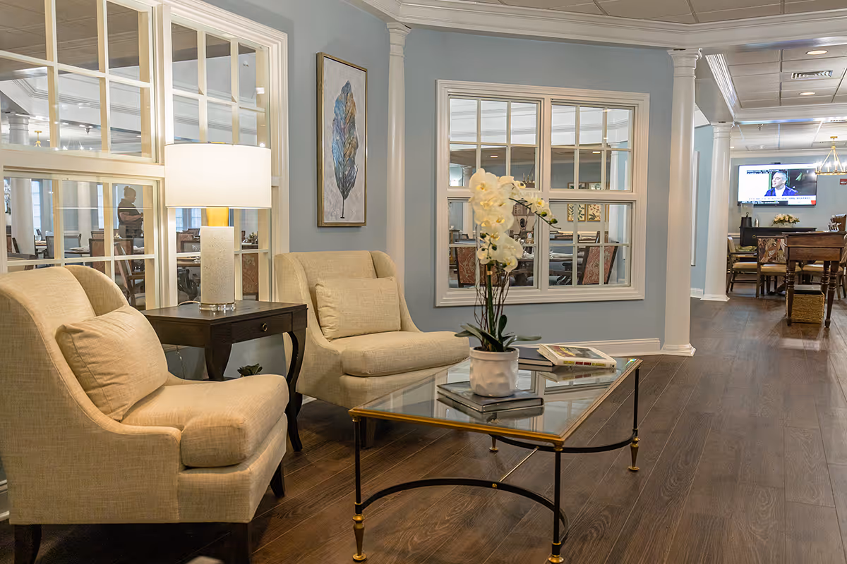 A cozy sitting area in a senior living facility with two beige armchairs, a glass coffee table with a potted orchid and books, a side table with a lamp, and large windows looking into an adjacent room with tables and chairs. The walls are painted light blue and decorated with a framed feather artwork.