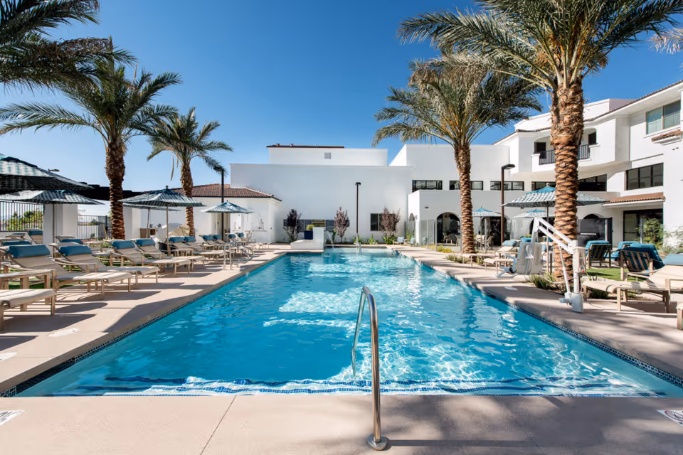Outdoor swimming pool at a senior living facility with lounge chairs and umbrellas along the sides, palm trees, and a clear blue sky. The building in the background is white with multiple windows and balconies.