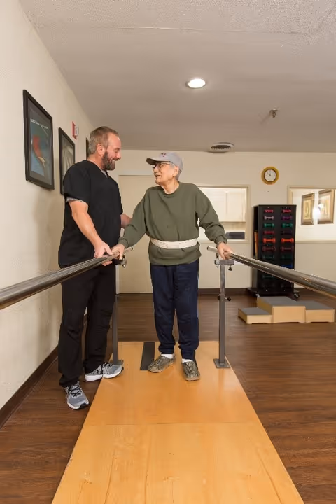 An elderly man wearing a green sweater, blue pants, and a gray cap is using parallel bars for walking exercise while being assisted by a man in black clothing. They are indoors in a room with wooden flooring and framed pictures on the wall.