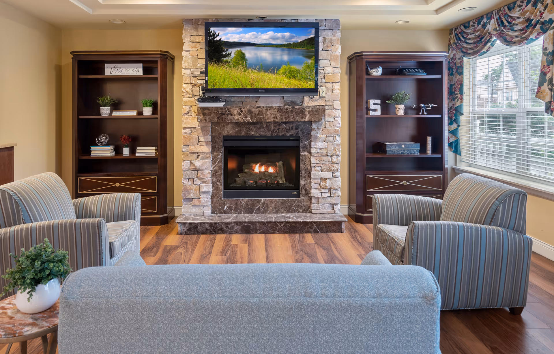 A cozy living room with a stone fireplace and a flat-screen TV mounted above it displaying a scenic lake view. The room features two dark wooden bookshelves on either side of the fireplace, decorated with plants, books, and small decorative items. There are two striped armchairs and a light blue sofa arranged around the fireplace on a wooden floor. A window with floral curtains allows natural light into the room.