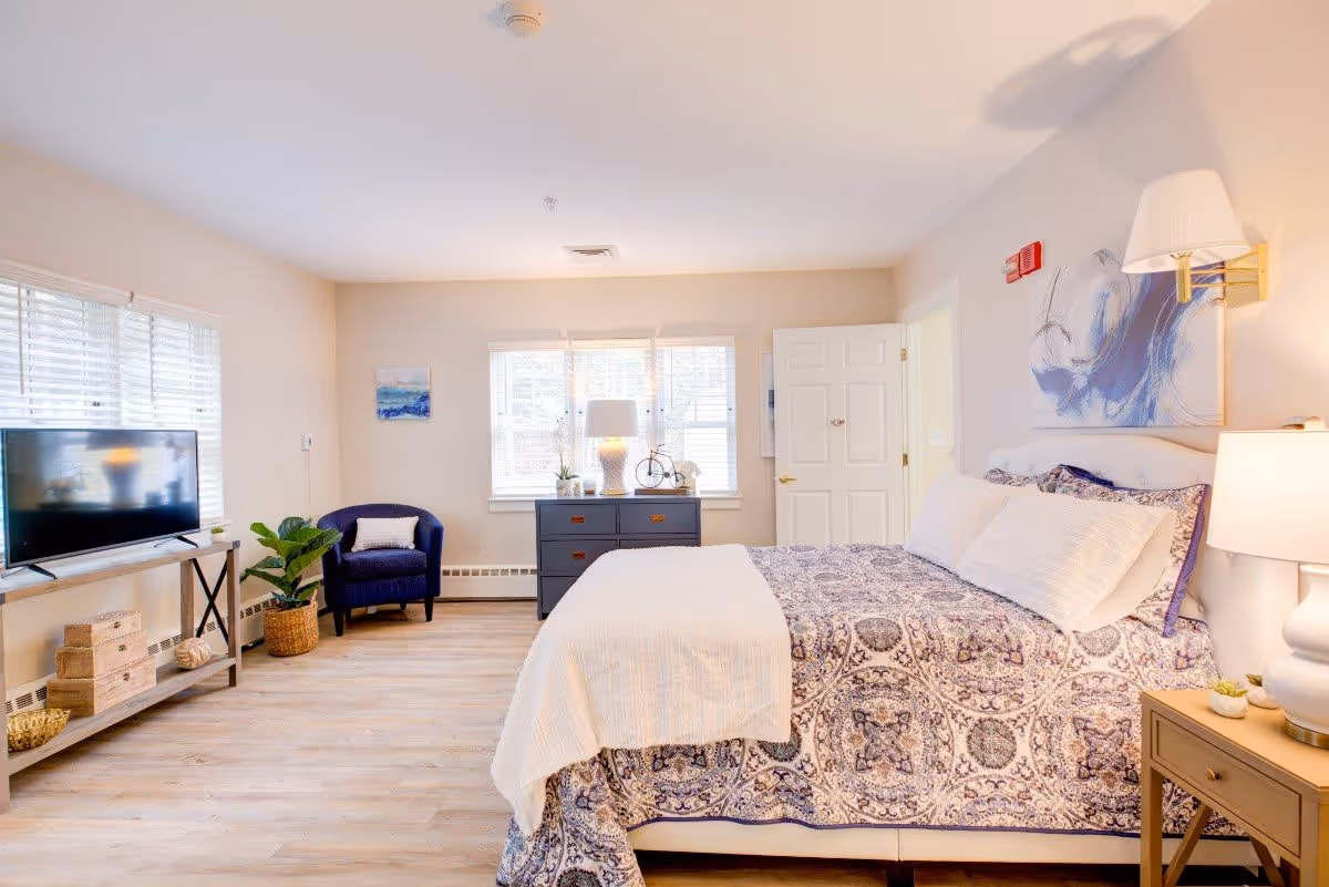 A bright and spacious bedroom with a large bed covered in a patterned purple and white bedspread. The room features light-colored walls and wood flooring. There is a blue armchair with a white pillow near a window with white blinds. A dark blue dresser with a lamp and decorative items sits under the window. A TV is placed on a wooden stand with shelves holding baskets and decor. A nightstand with a lamp and small plants is beside the bed. A door is open in the background, and a piece of abstract artwork hangs above the bed.