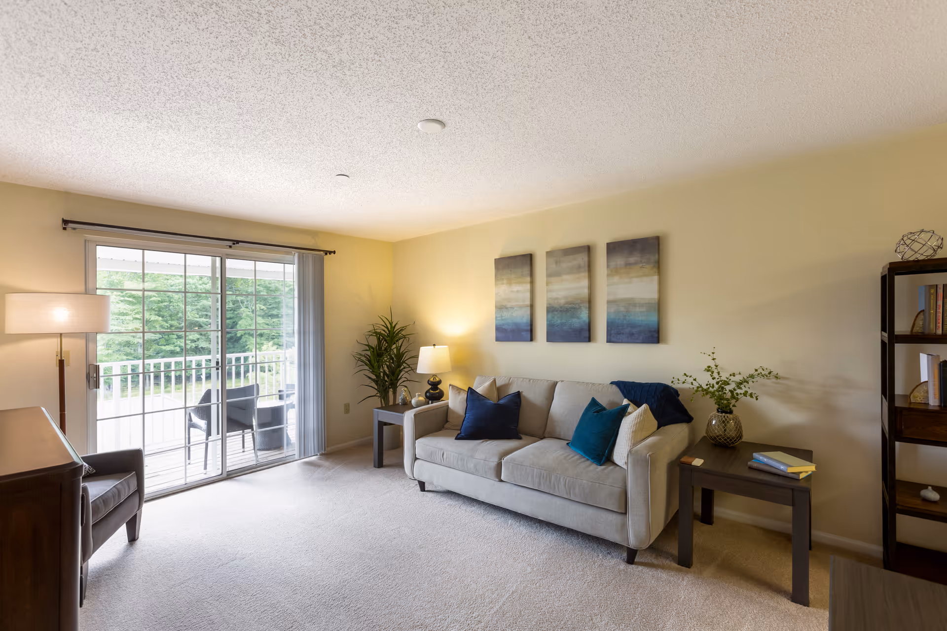 A cozy living room with a beige sofa adorned with blue and beige cushions, two side tables with lamps and a vase with greenery, a tall plant in the corner, a dark wooden bookshelf with books and decorative items, a sliding glass door leading to a balcony with outdoor chairs, and three abstract paintings on the wall above the sofa.