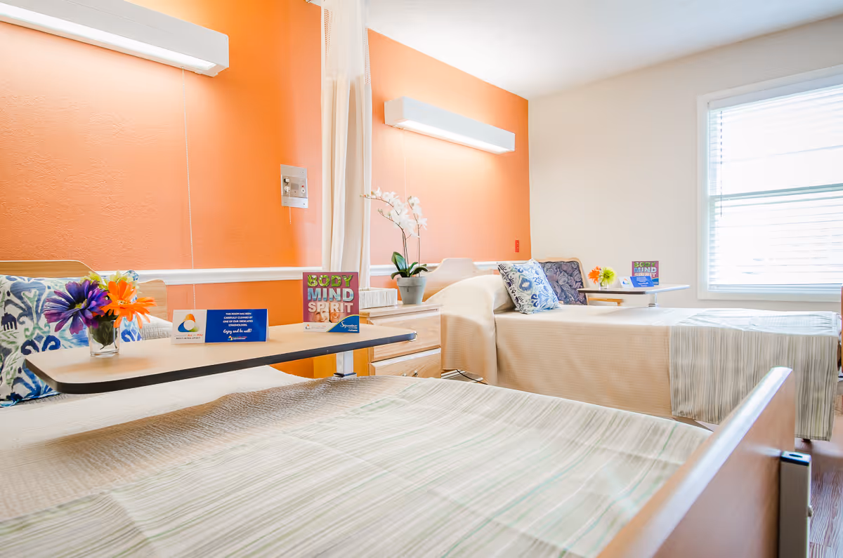 A bright and clean senior living bedroom with two beds, each with a small overbed table holding flowers and informational cards. The walls are painted orange and white, and there is a window with blinds letting in natural light. Decorative pillows and a potted orchid add a homey touch.