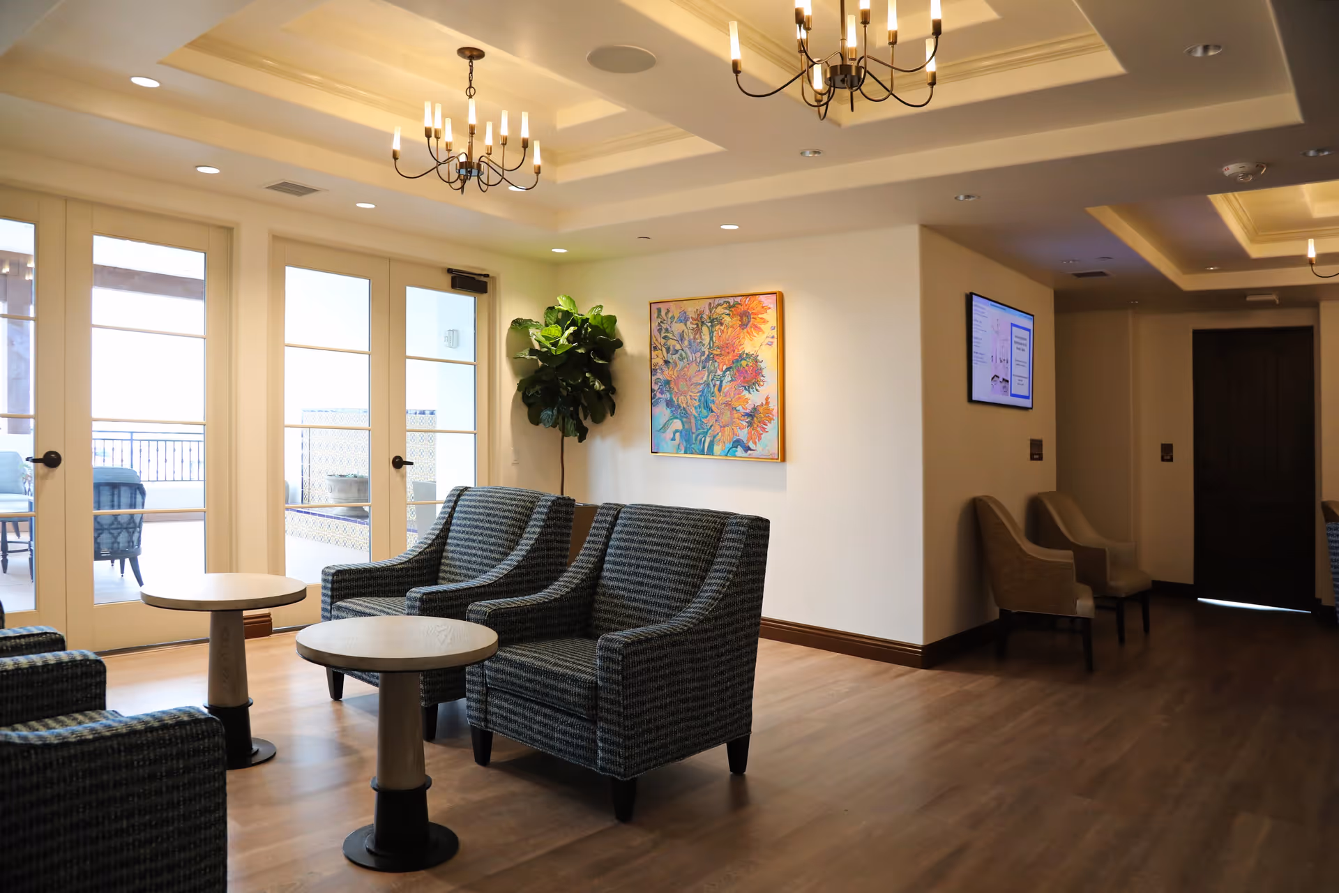 A bright and spacious sitting area with several upholstered armchairs arranged around small round tables. The room features large glass doors leading to an outdoor patio, a colorful floral painting on the wall, a tall green plant in the corner, and two chandeliers hanging from a coffered ceiling. There is also a wall-mounted screen and additional seating along the hallway.