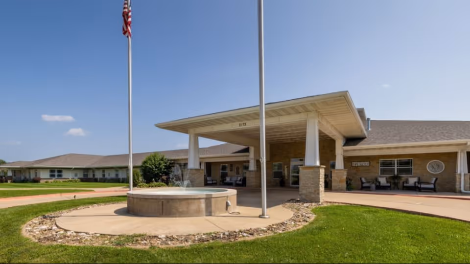 Front entrance of a senior living facility with a covered porte-cochere supported by columns, a circular fountain and flagpoles on a manicured lawn.