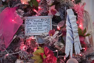 A close-up of a decorated Christmas tree featuring pink flowers, white gloves, and a small sign that reads 'Mothers hold their children's hands awhile and their hearts forever.' The tree is adorned with pink lights and frosted branches.
