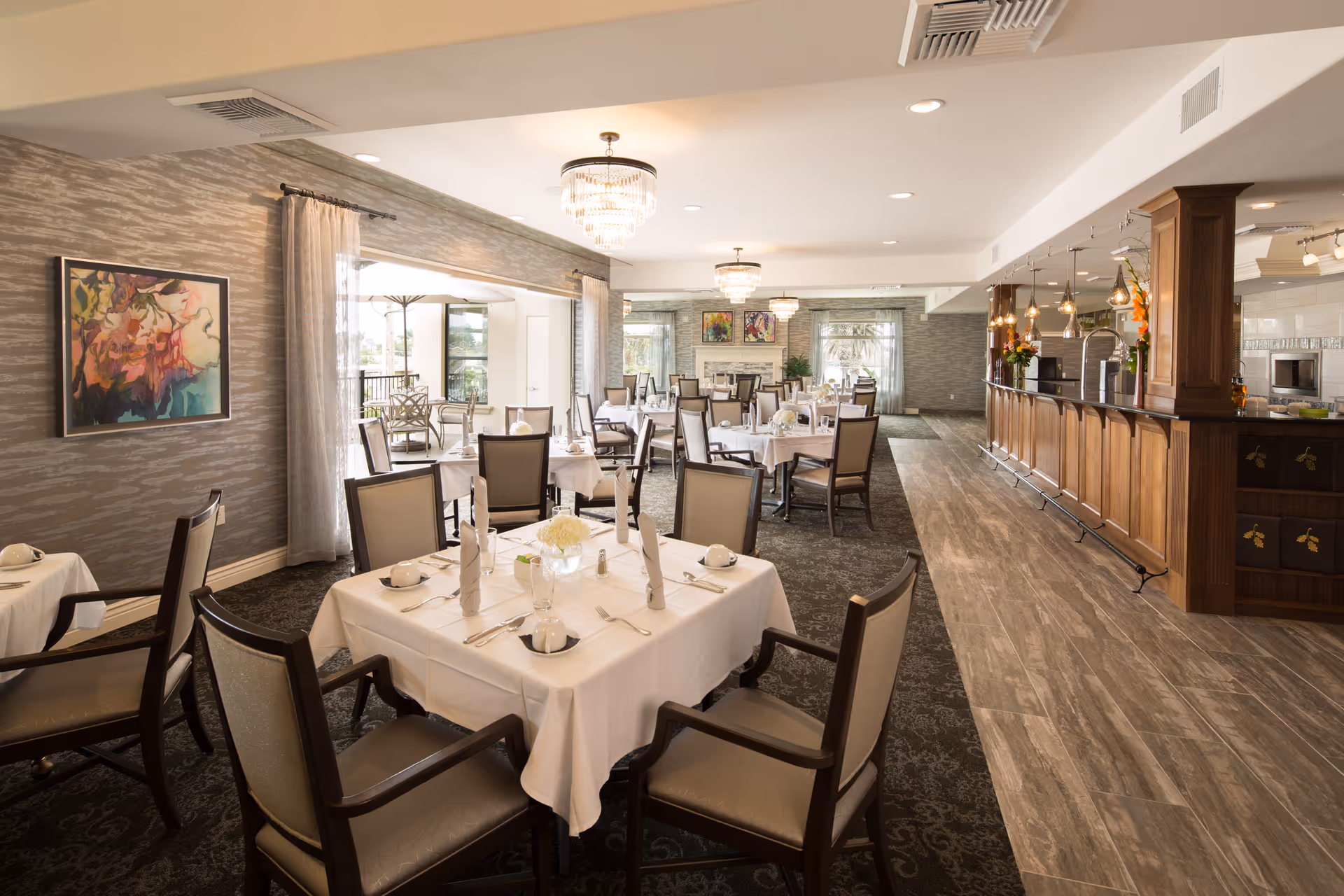 A spacious and elegant dining room with multiple tables covered in white tablecloths, each set with napkins, plates, and silverware. The room features comfortable chairs, a wooden bar area with hanging lights, large windows with curtains allowing natural light, and decorative artwork on the walls. The flooring is a combination of carpet and wood-like tiles.