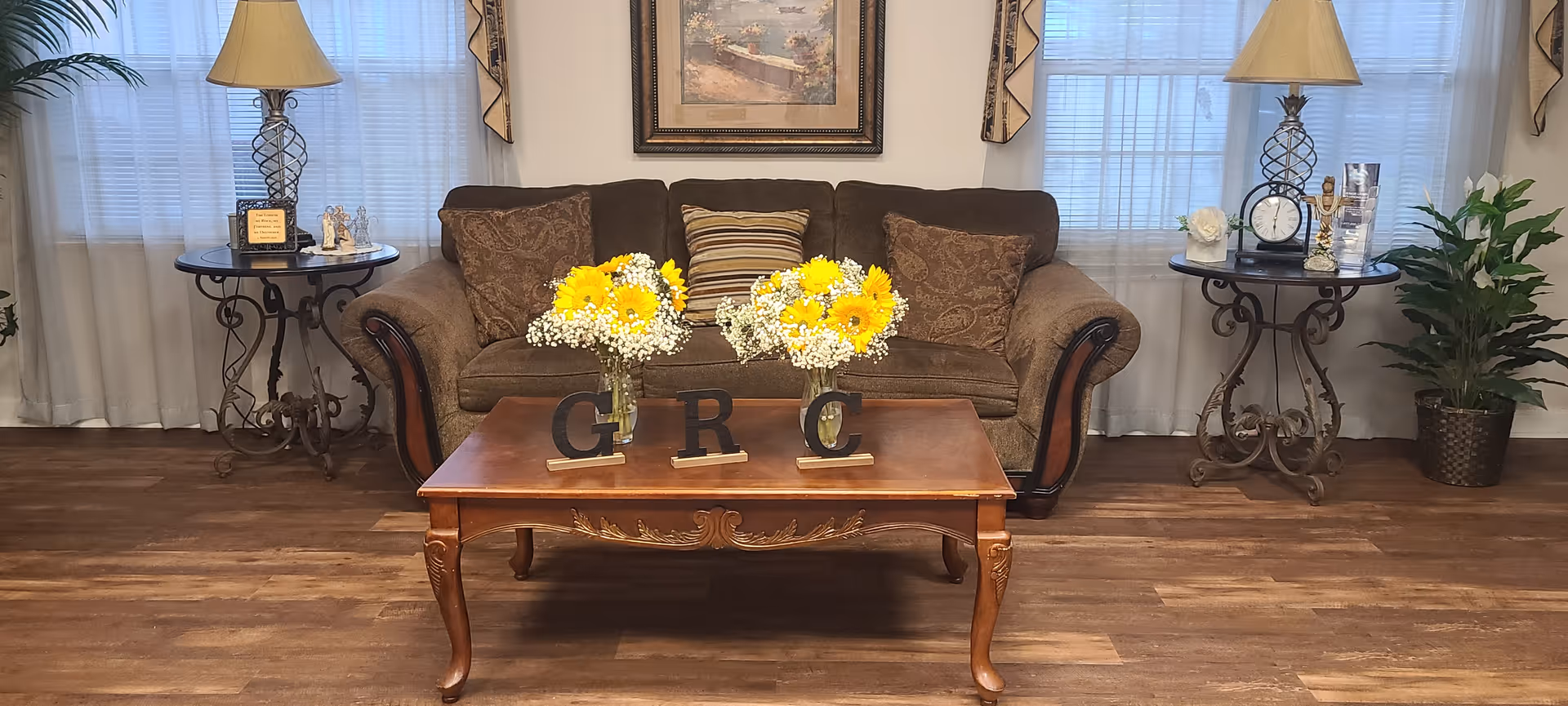 A cozy living room area with a brown sofa adorned with patterned and striped cushions. In front of the sofa is a wooden coffee table with two vases of yellow and white flowers and letters spelling 'G R C'. On either side of the sofa are round side tables with decorative lamps, small figurines, a clock, and other decor items. The room has wooden flooring, light-colored curtains, and a framed painting on the wall behind the sofa. A green potted plant is visible on the right side.