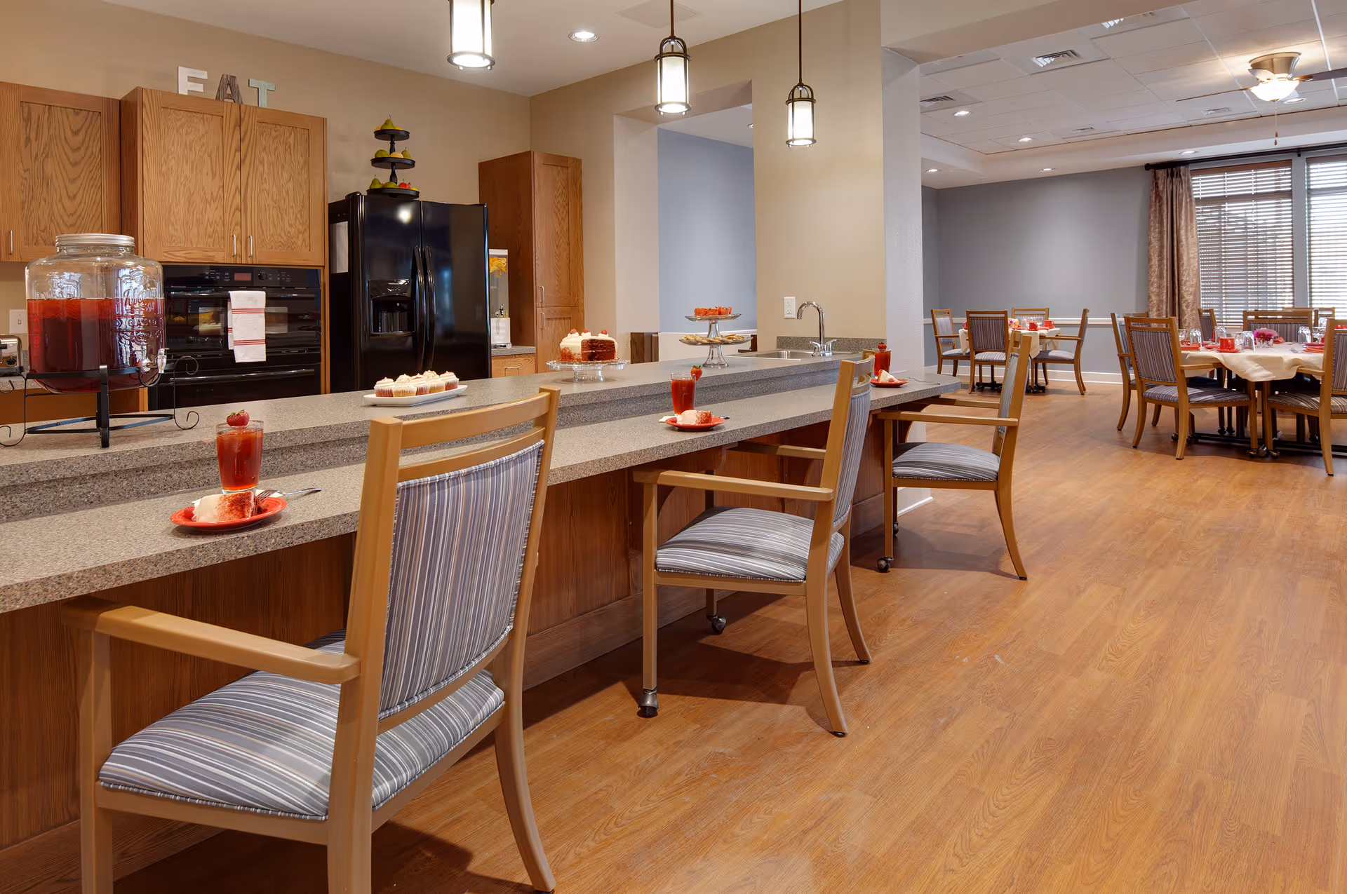 Communal dining area with a long serving counter and chairs set with drinks and desserts, and dining tables in the background.