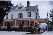Front exterior of a large white Victorian-style house with a wraparound porch decorated with string lights and snow on the ground.