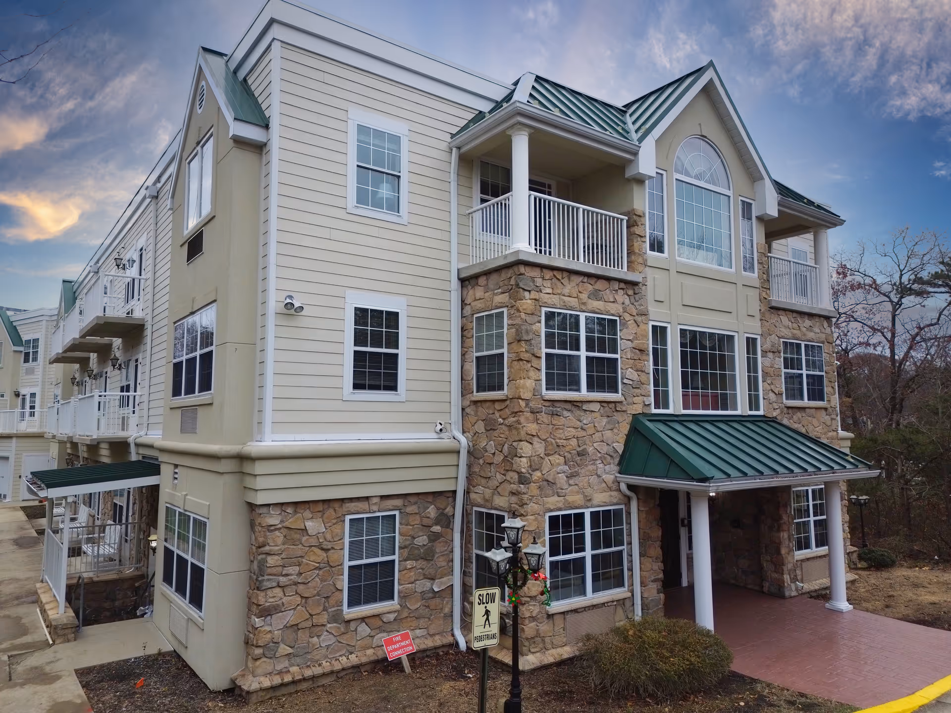Exterior view of a multi-story assisted living facility building with stone and beige siding, green metal roofs, multiple windows, balconies, and a covered entrance. The sky is partly cloudy with a sunset glow.
