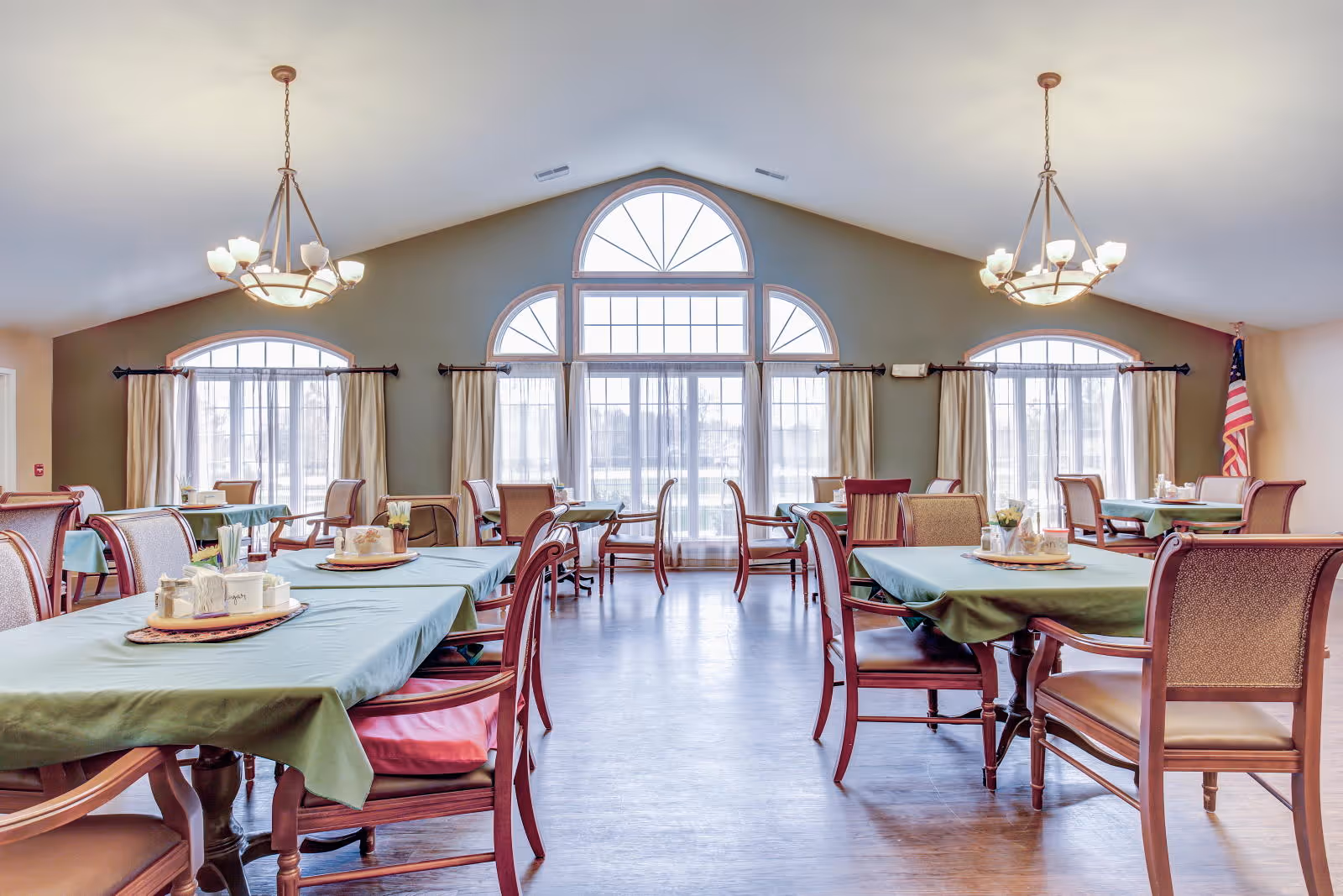 A bright dining room with multiple tables covered in green tablecloths and wooden chairs with cushions. The room features large arched windows with sheer curtains allowing natural light to fill the space. Two chandeliers hang from the ceiling, and an American flag is visible in the corner.