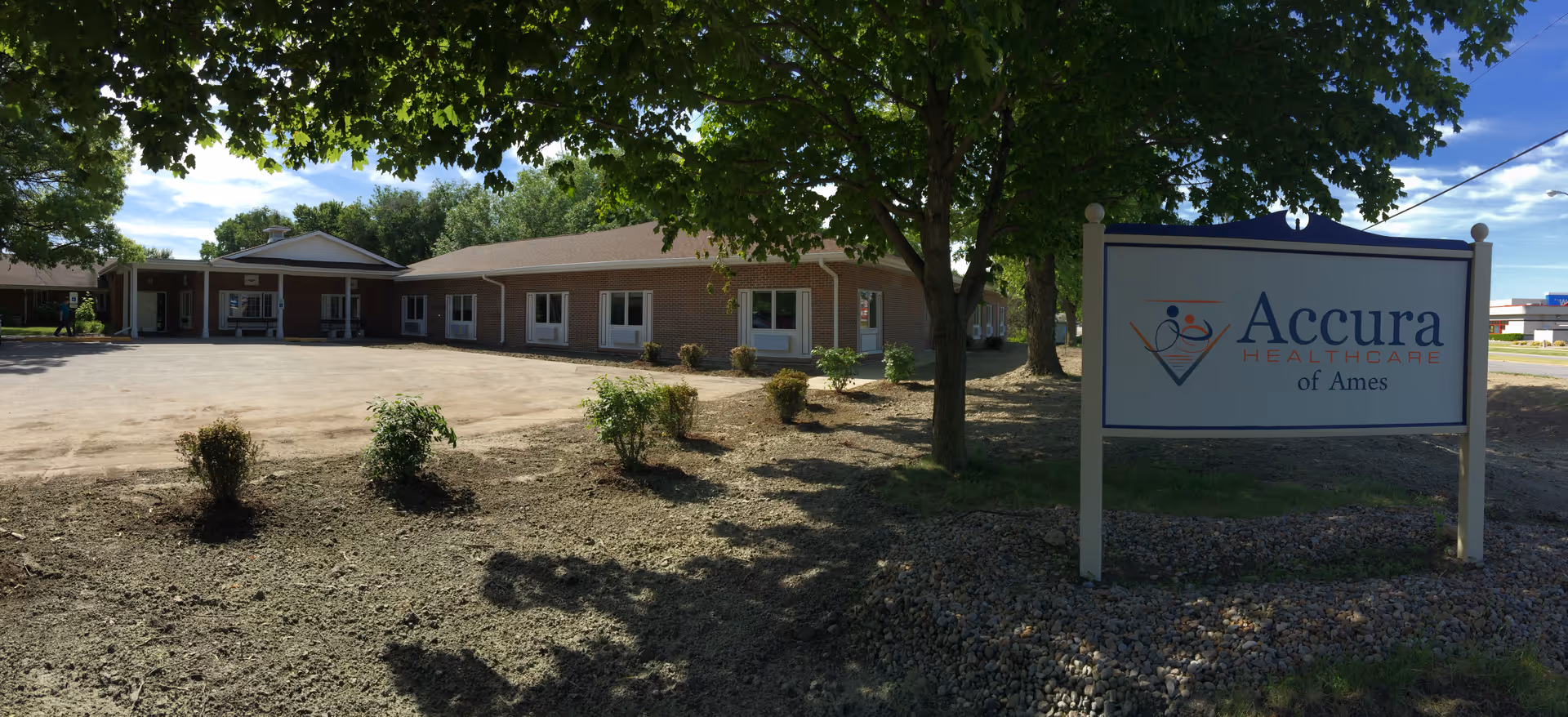 Exterior view of a single-story brick building with multiple windows, surrounded by trees and a dirt parking area. A large sign in the foreground reads 'Accura Healthcare of Ames'.