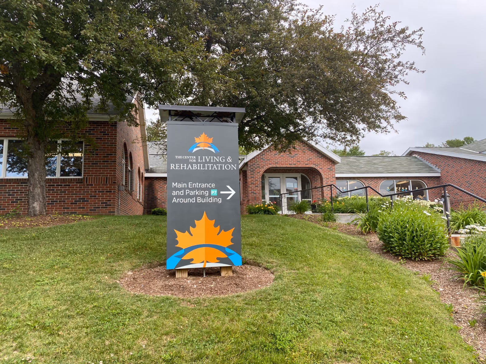Outdoor view of The Center for Living and Rehabilitation building with a sign in the foreground indicating the main entrance and parking are around the building. The building is made of red brick and surrounded by green grass, trees, and shrubs under a cloudy sky.