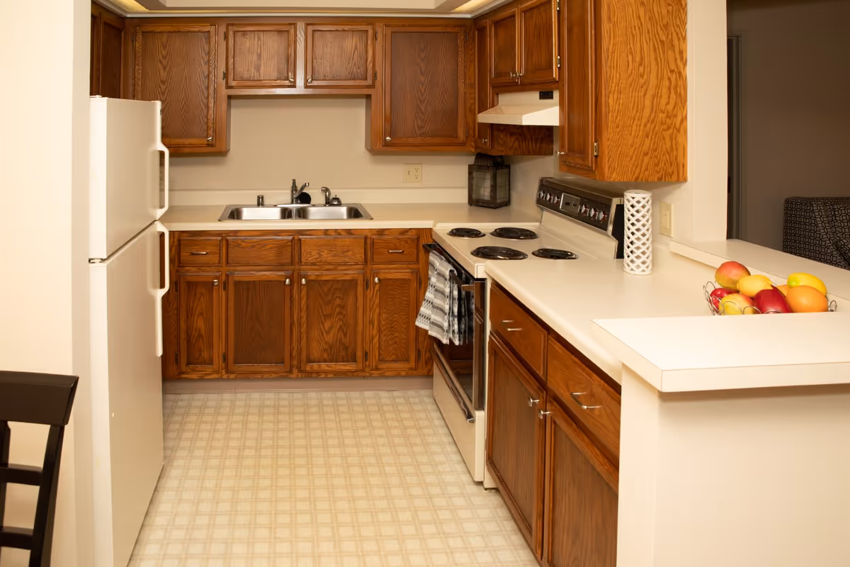 A mid-sized kitchen with oak cabinets, white countertops and appliances including a refrigerator, electric stove, double sink, and a bowl of fruit on the counter.