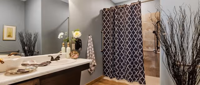 Bathroom with a shower area featuring a dark patterned shower curtain, a white sink countertop with soap dispensers and decorative items, a large mirror above the sink, a towel hanging on the wall, and tall decorative branches in a vase.