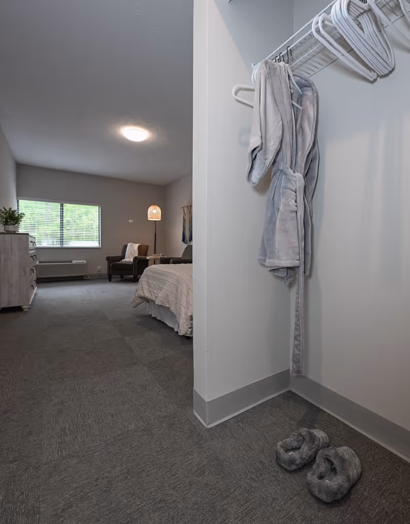 Carpeted senior living bedroom seen from an entry alcove with a gray robe hanging on a rack and fuzzy slippers on the floor in the foreground.