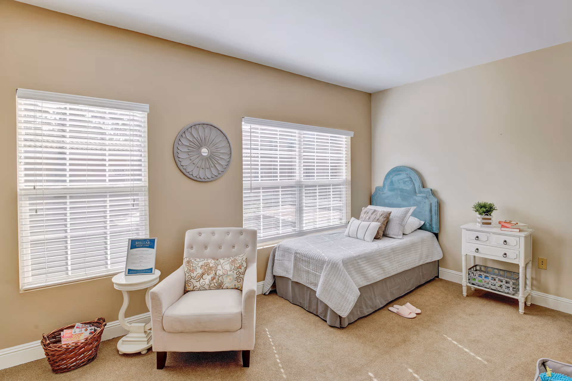 A cozy bedroom with a single bed featuring a blue headboard, white bedding, and multiple pillows. Next to the bed is a white nightstand with a small plant and books on top. There is a beige armchair with a decorative pillow and a small round white side table beside it. Two large windows with white blinds allow natural light to fill the room. A basket with magazines is placed on the floor near the chair.