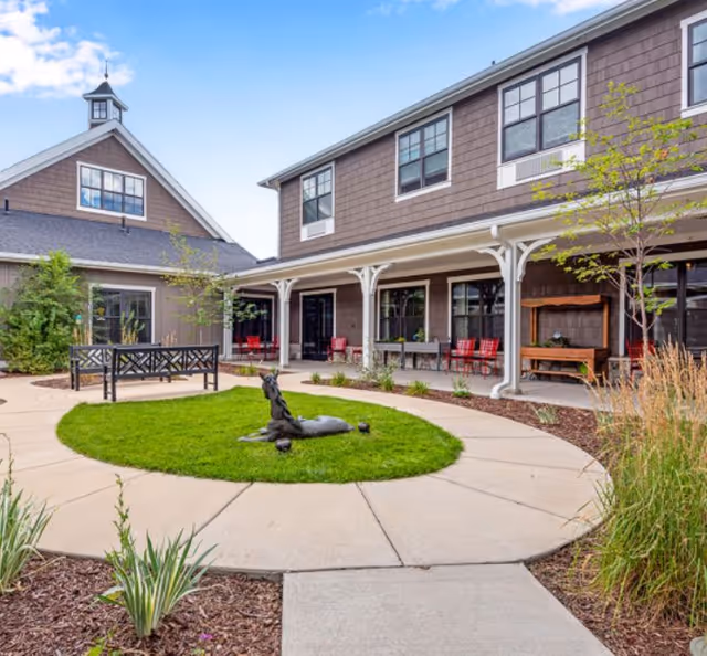 Sunny courtyard with a circular paved walkway around a grassy center featuring a bronze animal sculpture, benches, and a covered porch of a two-story building.