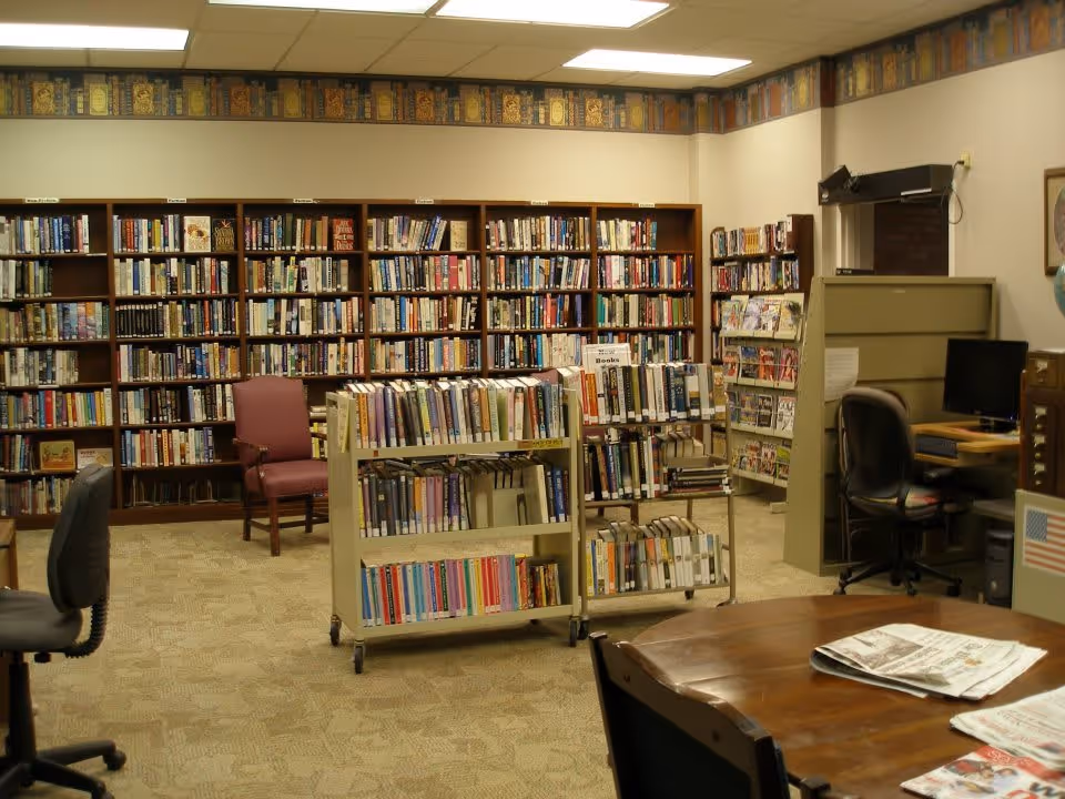 Interior library with bookshelves lining the wall, rolling book carts, chairs, a computer workstation, and a table with newspapers.