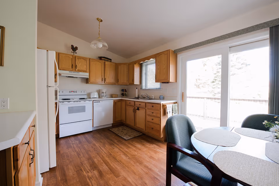 A bright kitchen with wooden cabinets, white appliances including a refrigerator, stove, and dishwasher, and a small dining area with a round table and chairs near a sliding glass door leading to an outdoor deck.