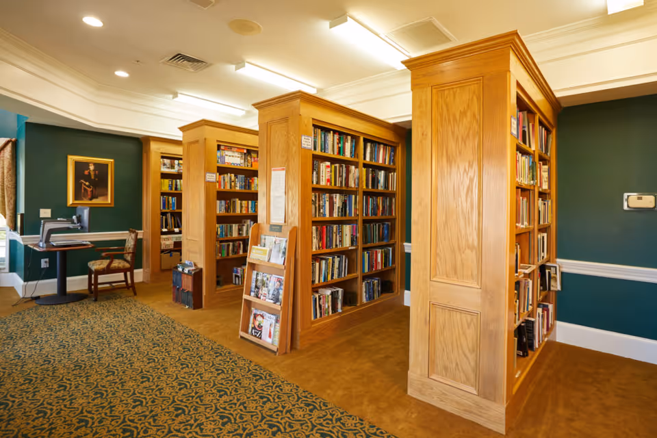 Interior view of a library room with wooden bookshelves filled with books. There is a patterned carpet on the floor, green walls with white trim, a framed portrait on the wall, and a computer workstation with a chair in the corner.