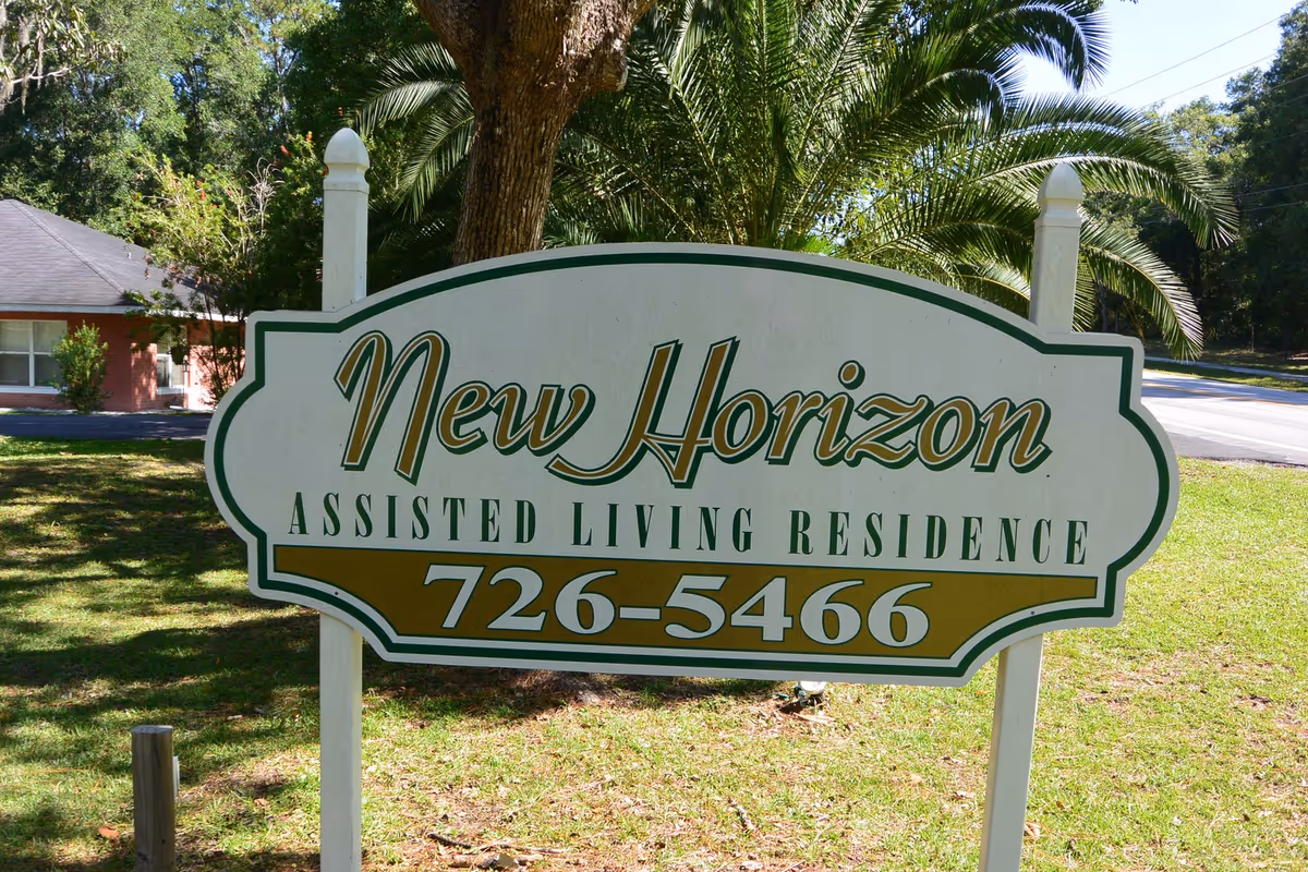 White and green sign reading "New Horizon Assisted Living Residence" and a phone number on a lawn with trees and a building in the background.
