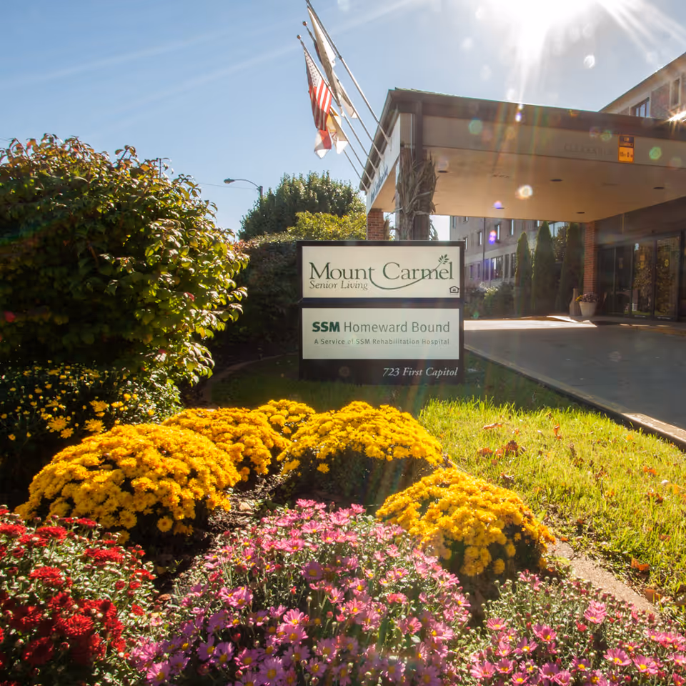 Entrance to Mount Carmel Senior Living facility with a sign displaying the name and SSM Homeward Bound service. The foreground features colorful flower beds with yellow, red, and pink flowers, and the building entrance is visible in the background under a canopy with flags flying above.