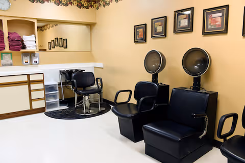 Interior view of a salon area in a senior living facility with black salon chairs, hair dryers, a styling chair, a large mirror, shelves with folded towels, and framed pictures on the wall.