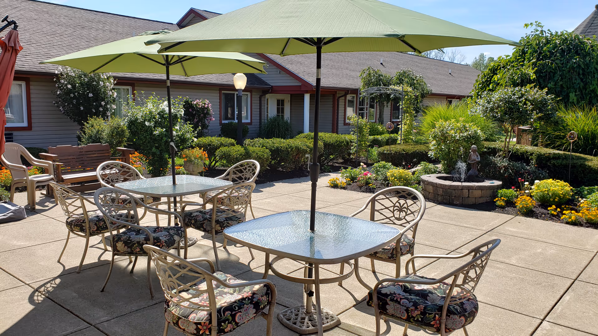 Sunny landscaped courtyard patio with glass-top tables, floral-cushioned chairs, large green umbrellas, and a small fountain in front of a single-story residential building.