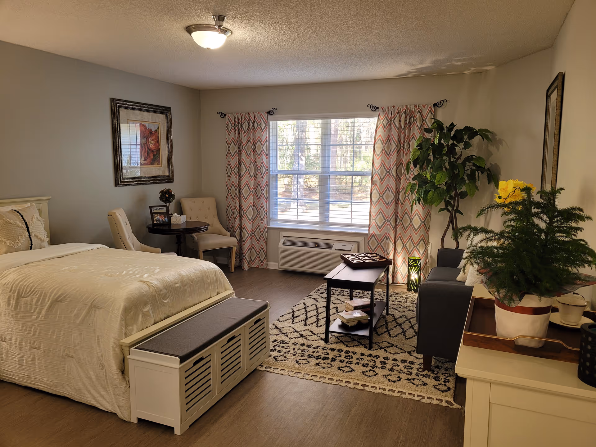 A cozy bedroom with a large bed covered in cream-colored bedding, a cushioned bench at the foot of the bed, two upholstered chairs with a small round table between them, a large window with patterned curtains, a gray sofa, a coffee table on a patterned rug, and several potted plants and framed artwork on the walls.