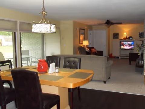 View of a combined dining and living room area in a senior living facility. The dining area features a wooden table with placemats, a red decorative jar, and dark upholstered chairs. Sliding glass doors with blinds lead to an outdoor patio with a table and chairs. The living room has beige sofas, a ceiling fan, a lamp, framed artwork on the walls, and a television displaying a colorful screen.