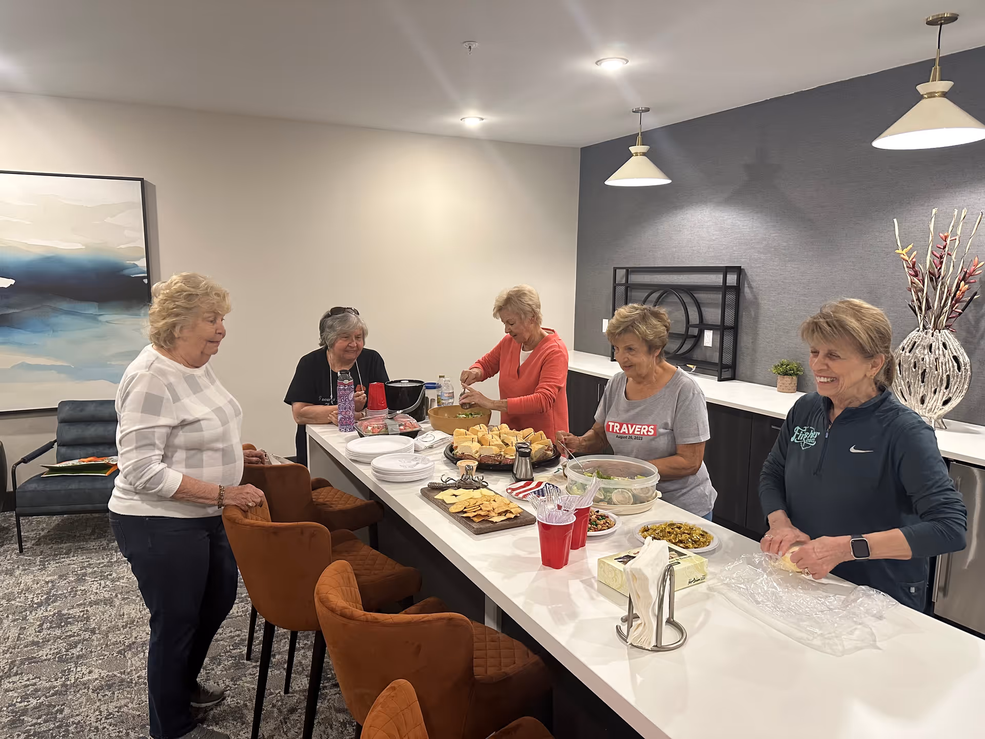 Five elderly women gathered around a kitchen island in a modern interior setting, preparing and serving food including bread, crackers, and salads. The room has contemporary decor with pendant lights, a large abstract painting, and comfortable seating.