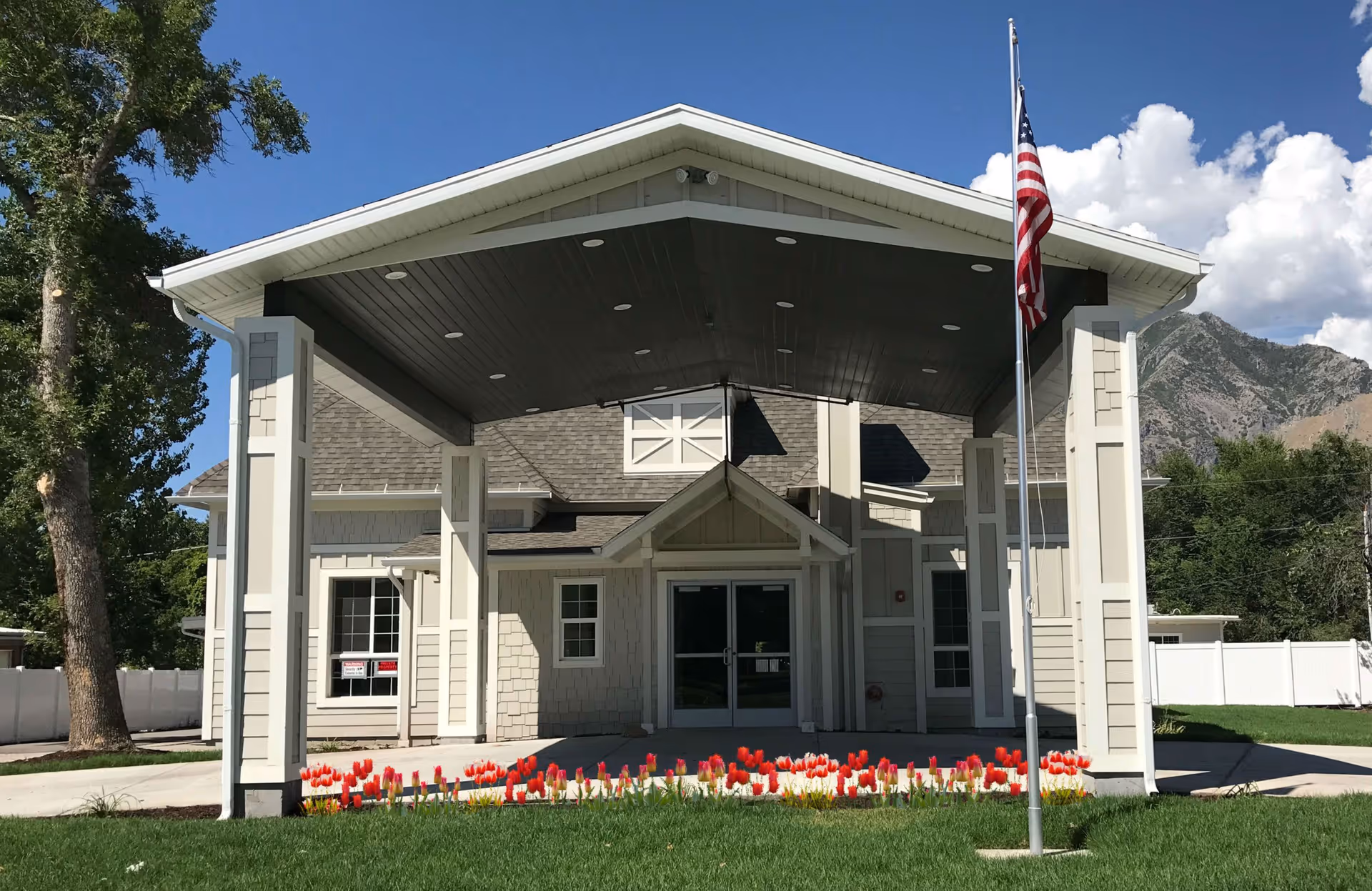 Front entrance of a beige assisted living building with a covered porte-cochère, American flag, and a bed of red tulips.