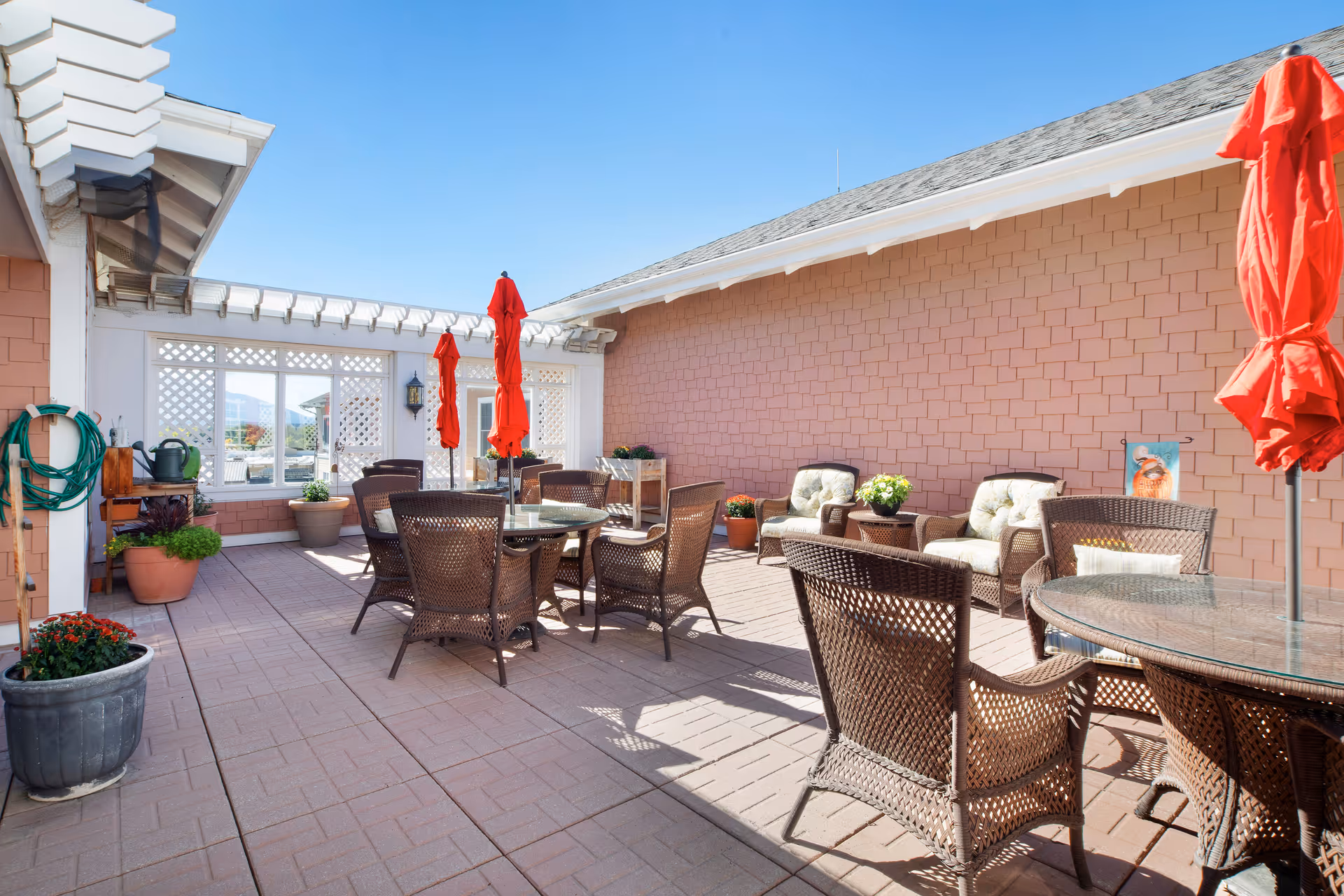 Outdoor patio area with multiple wicker chairs and glass-top tables, some with closed red umbrellas. The patio is surrounded by light brown shingle walls and decorated with potted plants. The sky is clear and blue.