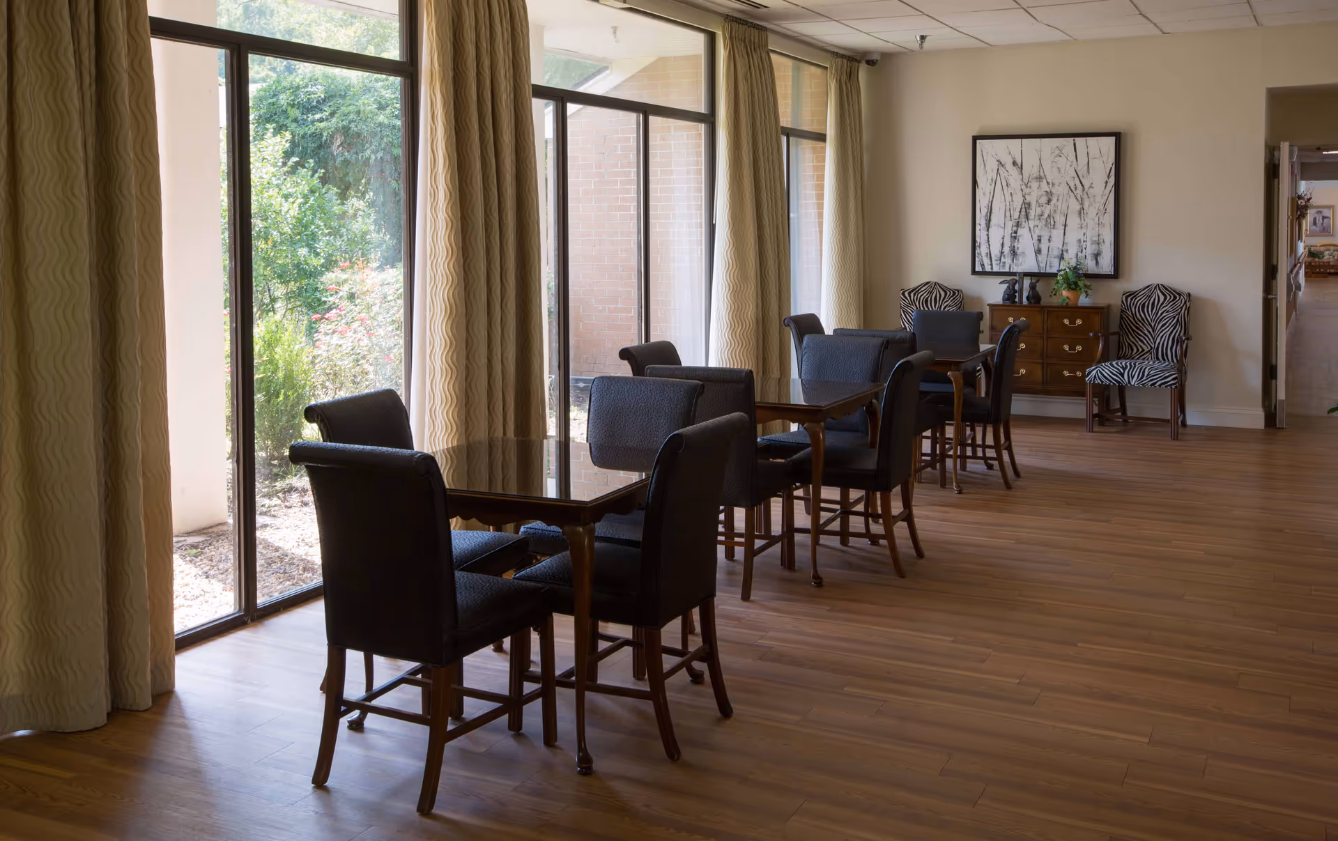 Sunlit communal dining area with several tables and upholstered chairs beside large windows and curtains.