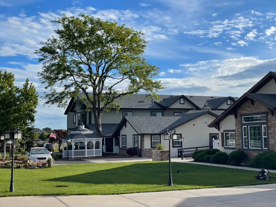 Exterior view of The Manor on Marston Lake facility showing a well-maintained lawn, a large tree, a white gazebo, several buildings with gray roofs and stone accents, a parked car, and a partly cloudy blue sky.