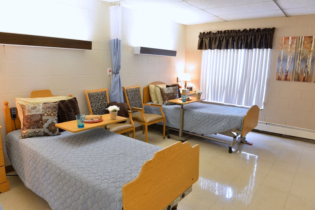 A bright room with two hospital-style beds covered with light blue quilts, each with a wooden headboard and footboard. Each bed has a small overbed table with a glass and a plate, and several decorative pillows. Between the beds are three wooden chairs with patterned cushions. A window with vertical blinds and a dark valance lets in natural light. A small lamp and a piece of artwork depicting birch trees are on the wall.