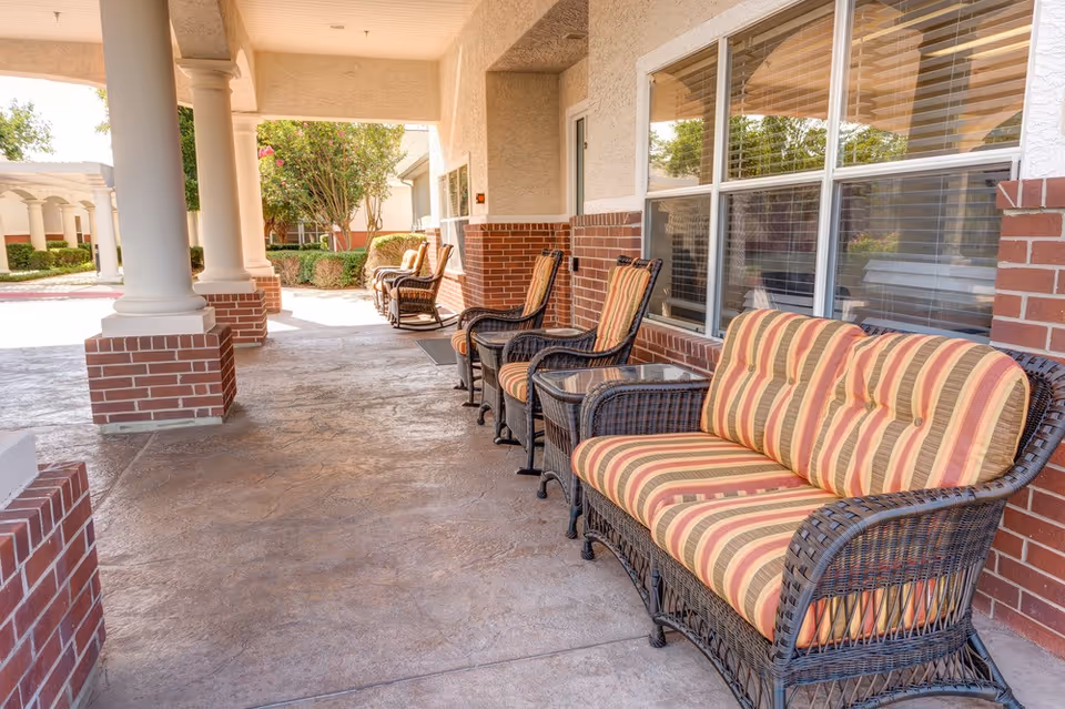Covered exterior porch with wicker chairs and striped cushions lined up along a brick wall and windows.