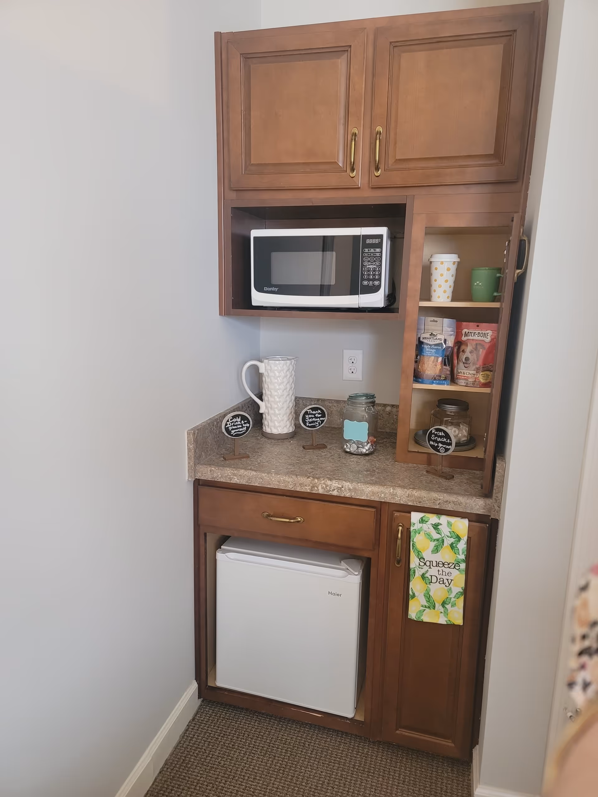 A small kitchenette area with wooden cabinets, a microwave, a mini refrigerator, and a countertop. On the countertop are a white pitcher, a glass jar with coins, and small signs indicating cold drinks, thanks for visiting, and fresh snacks. Inside an open cabinet are cups and snack bags. A towel with a lemon design and the words 'Squeeze the Day' hangs on the cabinet door.