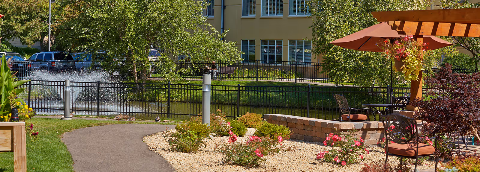 Outdoor garden area at Pearl Garden facility featuring a paved walking path, green grass, blooming flowers, a pond with a water fountain, metal fence, patio seating with cushioned chairs, a wooden pergola with a red umbrella, and trees providing shade.