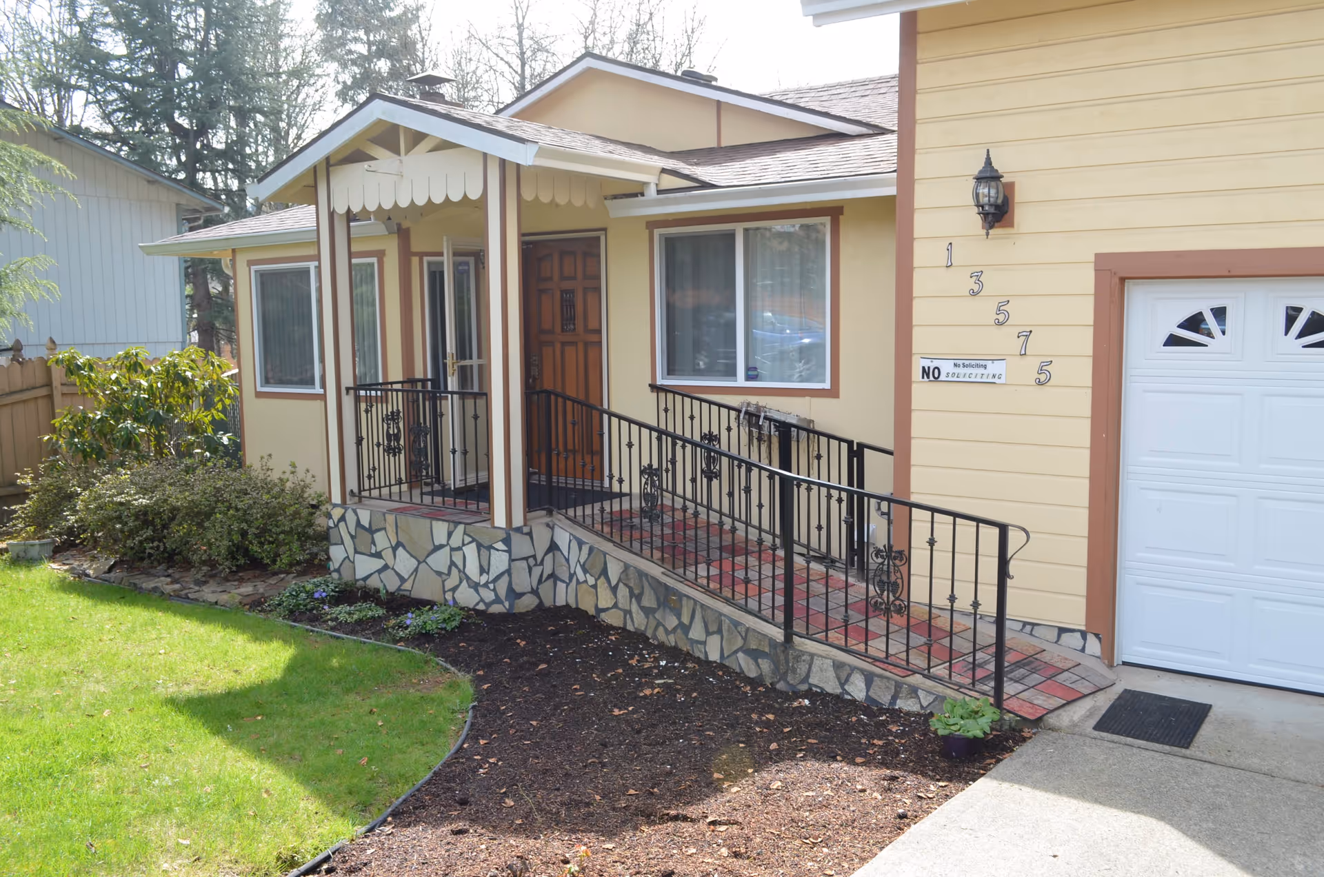 Front exterior view of a single-story yellow house with brown trim, featuring a wooden front door, a covered porch with a black metal railing and a wheelchair ramp, a white garage door, and a small garden with green grass and shrubs.