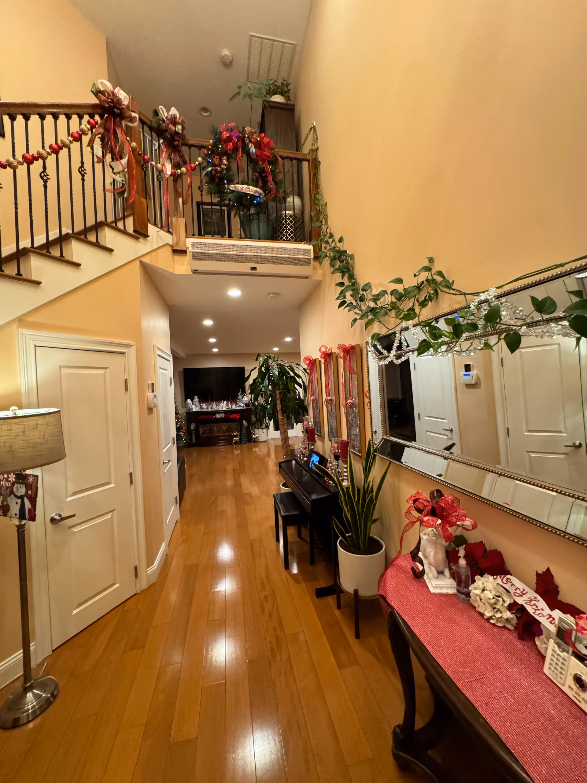 Interior hallway of a senior living facility decorated for Christmas with garlands, bows, and wreaths. The hallway features wooden flooring, a staircase with a decorated railing, a piano with a bench, plants, a large mirror on the wall, and a console table with holiday decorations. In the background, a living room area with a TV and more holiday decorations is visible.