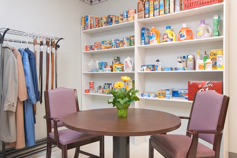 A small room with a round wooden table and two purple cushioned chairs. On the table is a green vase with yellow flowers. Behind the table are white shelves stocked with various household items including food products, cleaning supplies, and toiletries. To the left, there is a metal rack holding coats, shirts, and several belts.