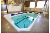 Indoor therapeutic pool with clear water, surrounded by tiled walls and large windows letting in natural light. The pool has handrails for easy access and steps leading into the water.