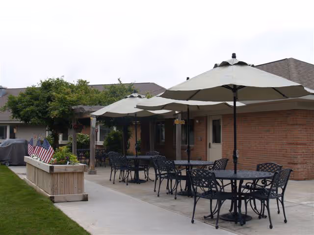 Outdoor patio area at Seminole Shores Living Center with several black metal tables and chairs under large beige umbrellas. The patio is adjacent to a brick building with doors and windows. There is a wooden planter box with American flags and greenery along the edge of the patio.