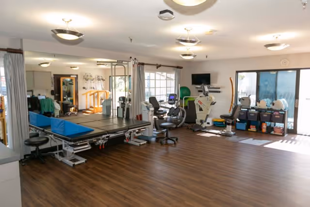 A spacious rehabilitation or physical therapy room with exercise equipment including stationary bikes, therapy tables, and various fitness and therapy tools. The room has large windows allowing natural light, wood flooring, and ceiling lights.
